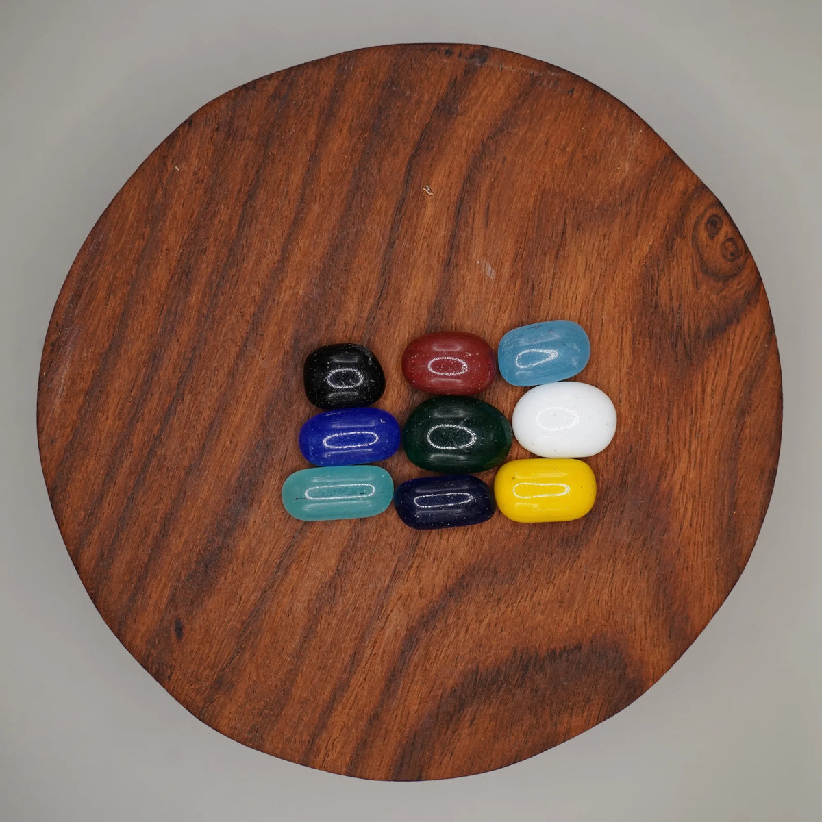 Nine smooth coloured puja stones in black, maroon, blue, green, white and yellow on a round wooden plate