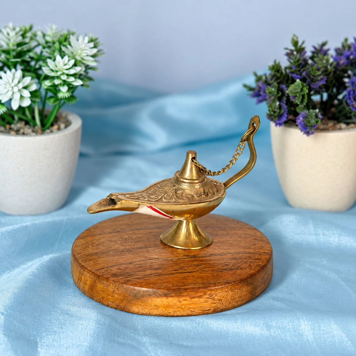 Brass Aladdin-style chirag lamp with an engraved body, a lid on a chain, a curved handle and a wick in the spout, on a round wooden base