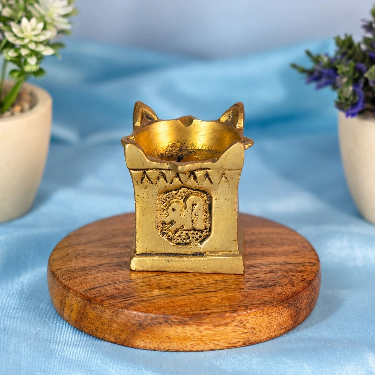 Small brass tulsi diya shaped like a square platform with corner horns, a Shri emblem on the front panel and a round oil bowl on top, on a round wooden base