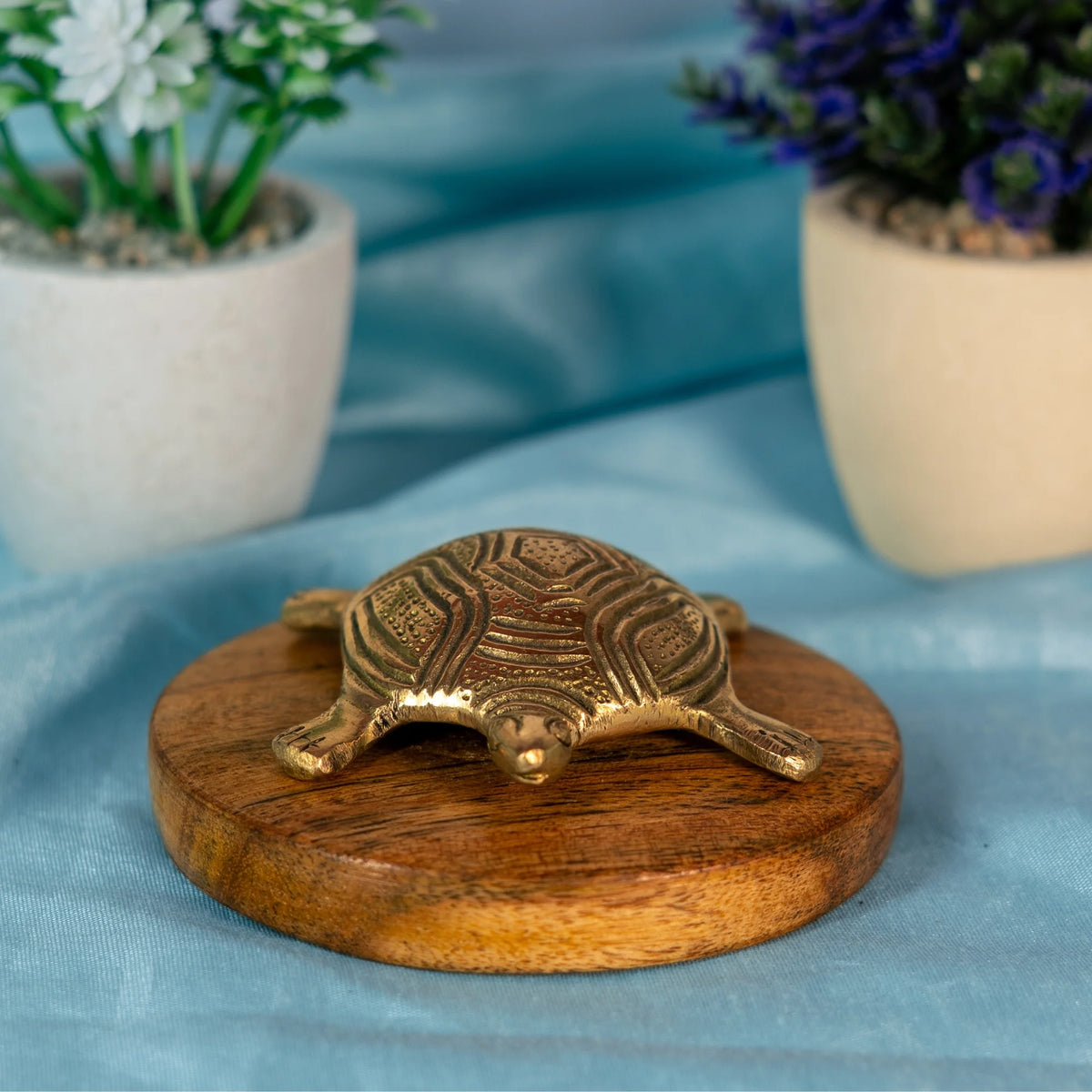 Small brass tortoise with a shell carved in geometric panels and curved bands, head forward and legs spread, on a round wooden base with a blue cloth behind