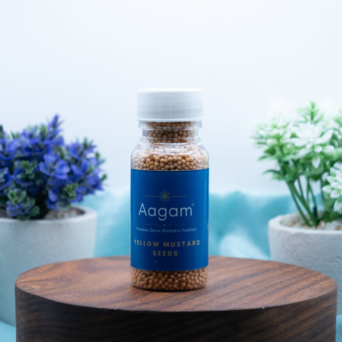 Aagam yellow mustard seeds in a clear bottle with a blue label and white cap, filled with golden-yellow seeds, shown on a wooden stand